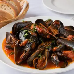 Mejillones a la gallega servidos en un plato blanco con salsa de tomate y perejil picado. Al fondo, una cesta de pan y una copa de vino.