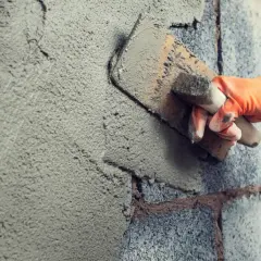 Mano con guanto arancione stende malta per intonaco esterno su un muro di blocchi di cemento con una cazzuola.