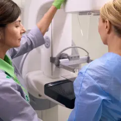 Lekarka w rękawiczce przygotowuje pacjentkę do badania mammograficznego. Widoczny jest fragment aparatu i pierś, czyli sutek, który będzie badany.