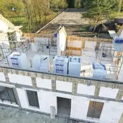 Budowa domu w toku, widoczne szalunki i materiały budowlane. Widać charakterystyczne trójkątne **goble** dachu, które czekają na pokrycie.