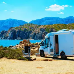 Un camping-car blanc est garé sur un terrain sablonneux, face à une mer bleue et des montagnes. L'application spot camping car idéale pour ce paysage.