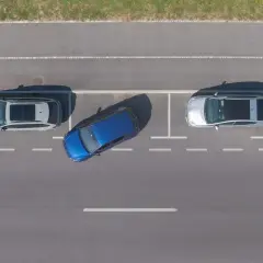 Niebieski samochód wykonuje parkowanie równoległe tyłem między dwoma innymi autami na parkingu.