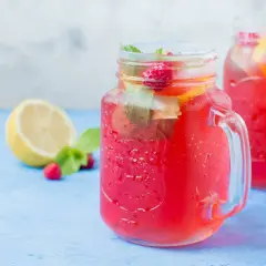 Orzeźwiająca lemoniada malinowa w słoikach z lodem, malinami i miętą. Obok połówka cytryny.