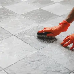 Mani con guanti arancioni spazzolano le fughe del pavimento con schiuma, mostrando come pulire le fughe del pavimento per un risultato brillante.