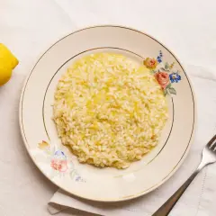 Un cremoso risotto al limon, con ralladura de limón espolvoreada, servido en un plato floral. Dos limones frescos reposan cerca.