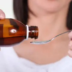 Mano versa sciroppo da una bottiglia marrone su un cucchiaino. Un rimedio per la tosse, un sedativo tosse, pronto per essere assunto.