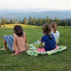 Trzy dziewczynki, jak małe rysianki, siedzą na trawie, podziwiając widok gór. Jedna z nich, z żabnicą na głowie, wpatruje się w dal.