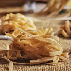 Nidi di tagliatelle fatte in casa, pronte per essere cucinate. La pasta fresca, con la sua texture rustica, è disposta su una tela grezza, invitando a un pasto autentico.