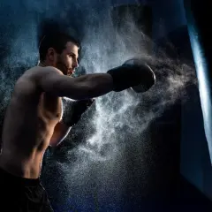 A determined boxer, embodying the characteristics of a fighter, throws a punch at a heavy bag, kicking up a cloud of dust.