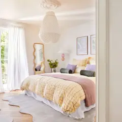 Ein geschwungener Spiegel im Schlafzimmer reflektiert das helle Zimmer mit Bett, weißen Vorhängen und Blick ins Grüne.