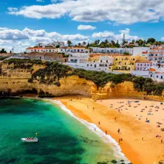 Sonnige Küste Portugals im Oktober. Ein Boot auf dem türkisfarbenen Meer vor einer Bucht mit Sandstrand und weiß-gelben Häusern.