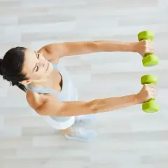 Kobieta wykonuje ćwiczenia z hantlami w domu, unosząc je przed sobą.