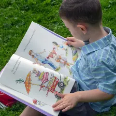 Chłopiec w niebieskiej koszuli czyta otwartą książkę z kolorowymi ilustracjami na trawie.