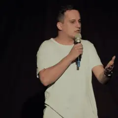 Mężczyzna w białej koszulce stoi na scenie z mikrofonem w dłoni, gestykulując.