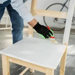 Persona con guanti verdi e neri sta per verniciare mobile legno con vernice bianca, usando un pennello.