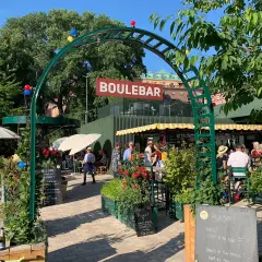 Sommar vid boulebar Rålambshov. Folk sitter vid bord under gula parasoller, grönska och röda blommor.