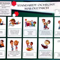 Tablica z ważnymi numerami telefonów i standardami ochrony dzieci. Widać rysunki dzieci, np. bawiące się, uczące, pomagające sobie. Reagujemy na każdy sygnał przemocy.