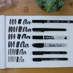 Práctica de lettering con varios rotuladores, mostrando trazos y letras "m" y "a" en un cuaderno.