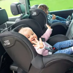 Uśmiechnięte niemowlę w foteliku samochodowym Britax Römer. Obok starsze dziecko. Bezpieczna podróż do jakiego wieku dziecko w foteliku jest kluczowa.