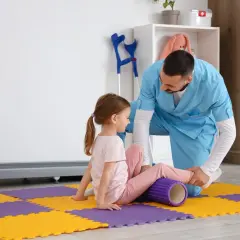 Fizjoterapeuta stosuje metodę Bobath podczas ćwiczeń z dziewczynką, która leży na macie z wałkiem pod nogą.