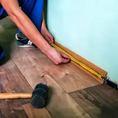 Mężczyzna w niebieskich spodniach mierzy taśmą podłogę, przygotowując się do tego, jak kłaść panele. Obok leży gumowy młotek.