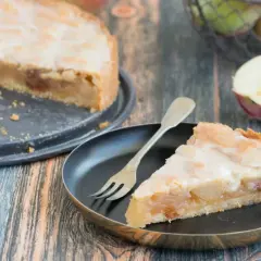 Ein Stück von einem köstlichen gedeckten Apfelkuchen mit süßer Glasur auf einem Teller, daneben ein ganzer Kuchen und frische Äpfel.