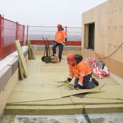 Dwóch budowlańców w pomarańczowych kurtkach pracuje na budowie. Układają płyty izolacyjne na płaskim dachu.