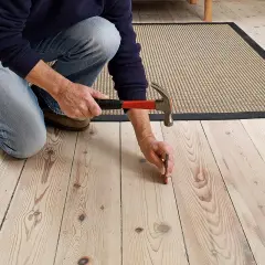 Person knäböjer och slår en spik i ett trägolv. En matta ligger bredvid.