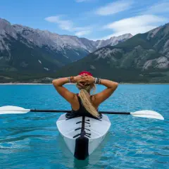 Frau im Kajak auf türkisfarbenem See, Berge im Hintergrund. Ein Symbol für das Erreichen von finanzieller Freiheit und ein Leben voller Abenteuer.