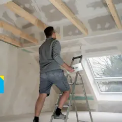 Un operaio su una scala lavora al soffitto, applicando stucco su pannelli di fibrogesso, materiale versatile per ristrutturazioni.