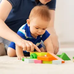 Dziecko w niebieskiej koszulce bawi się kolorowymi klockami na dywanie, z pomocą dorosłego.