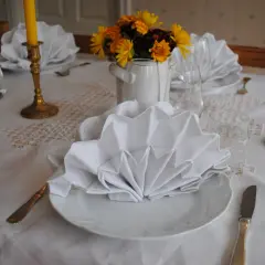 Vackert dukat bord med vita, vikta servetter i glas. Guldljusstakar och blommor skapar en festlig stämning.