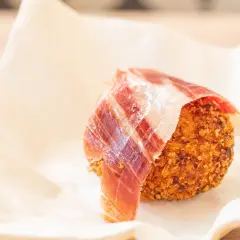 Croquetas de jamón crujientes, envueltas en una loncha de jamón serrano, servidas sobre papel blanco.