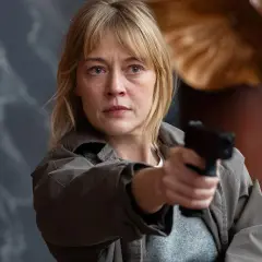 En kvinna med blont hår och en grå jacka håller en pistol redo. Hon är en del av rollistan i morden och ser beslutsam ut.