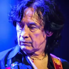 Andrzej Łabędzki z Lady Pank na koncercie, w niebieskim świetle, z charakterystyczną fryzurą i naszyjnikiem.