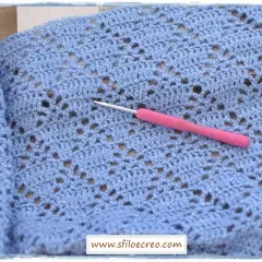 Un uncinetto rosa riposa su un lavoro a maglia azzurro con fili scintillanti, mostrando un delicato punto incrociato uncinetto.