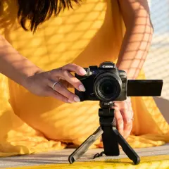 Kobieta w żółtej sukience ustawia aparat Canon EOS R10 na statywie, eksperymentując z trybem manualnym w aparacie.