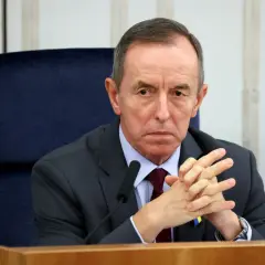 Tomasz Grodzki w garniturze, z rękami złożonymi na piersi, siedzi przy stole. Na klapie marynarki przypinka Poczty Polskiej.