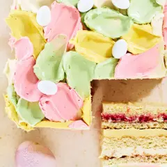 En vacker vit choklad tårta med pastellfärgad frosting och påskägg. En bit är uppskuren och visar lager av kräm och sylt.