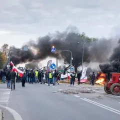 Czerwony traktor na drodze, w tle dym i ludzie z polskimi flagami.
