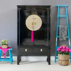 Mobile con laccatura nera lucida, dettagli dorati e nappa fucsia. Accanto, sgabello blu con fiori e scaletta decorativa.