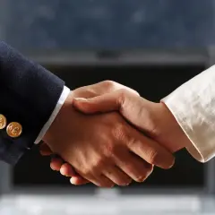 Uścisk dłoni symbolizujący udane firmanctwo. Dwie osoby w eleganckich strojach, w tle laptop.