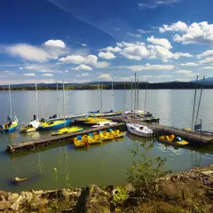 Żaglówki i rowerki wodne cumują przy pomoście na Jeziorze Żywieckim. Błękitne niebo z chmurami i spokojna tafla wody.