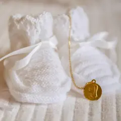 Douces chaussons blancs tricotés, ornés de rubans, avec une médaille dorée de la Vierge. Un symbole de marraine sans baptême, plein d'amour.