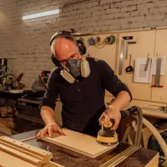 Artisan concentré, masqué et casqué, réalisant un ponçage bois méticuleux avec une ponceuse électrique dans son atelier.