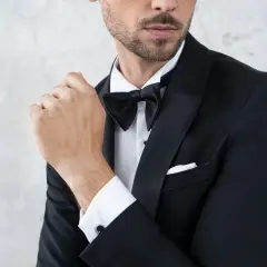 Elegancki mężczyzna w czarnym smokingu i muszce, z dłonią przy ustach.