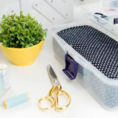 Un angolo del craft room con forbici dorate, rocchetti di filo azzurro, un vaso giallo con una pianta e una scatola trasparente con stoffa a pois.