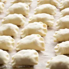Gotowe do gotowania pierogi, przygotowane według tradycyjnego przepisu. Widać je ułożone rzędami na blacie.