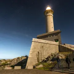 El faro de Punta Nariga ilumina la noche estrellada. Tres personas suben las escaleras hacia la torre, bajo un cielo lleno de estrellas.