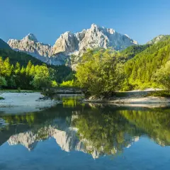 En vacker vy för att vandra i Slovenien. En spegelblank sjö reflekterar berg och skog. En bro leder över vattnet.
